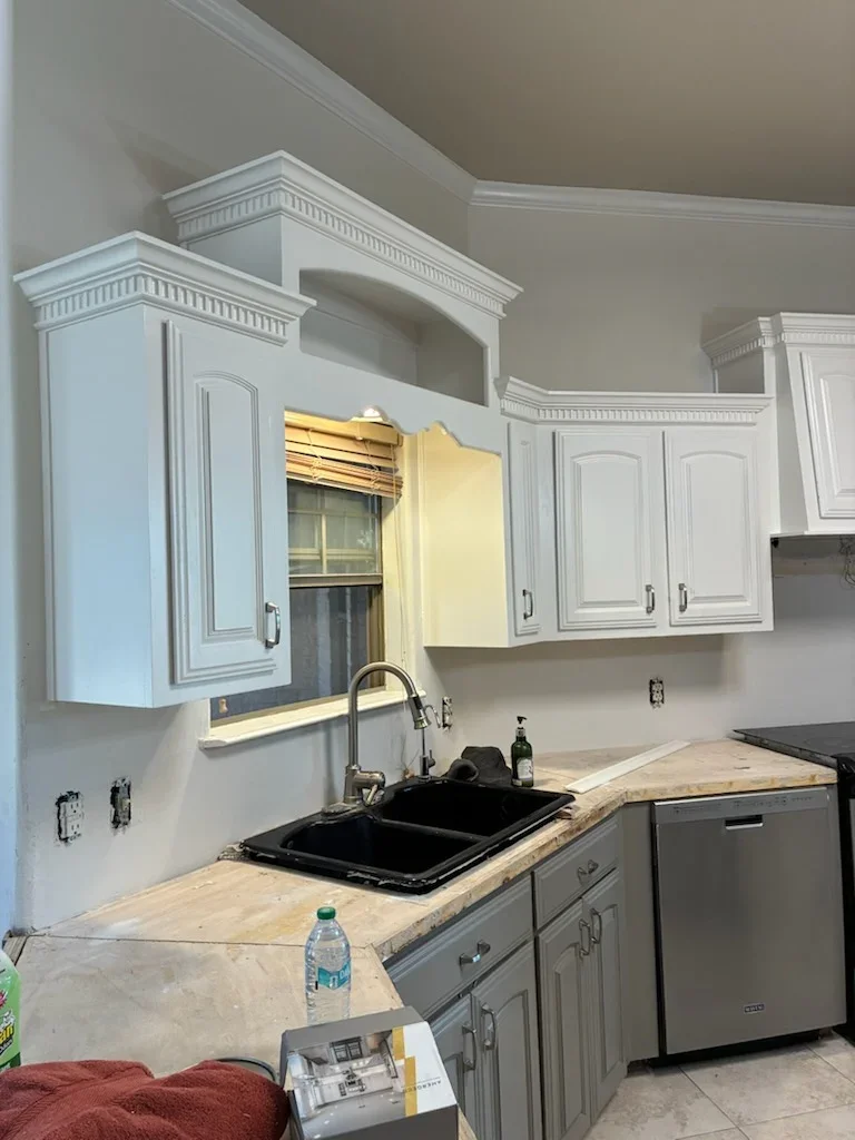 White painted kitchen cabinets with crown molding, neutral walls, black sink, and light countertops