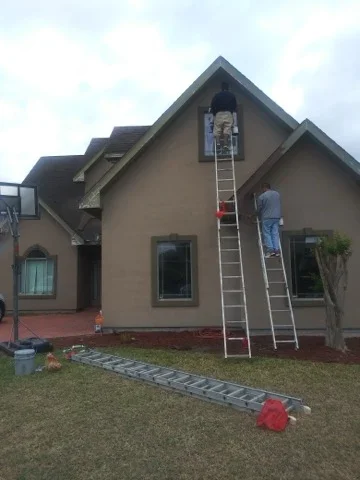 Exterior house painting with tan stucco and dark trim, ladders and protective coverings in place