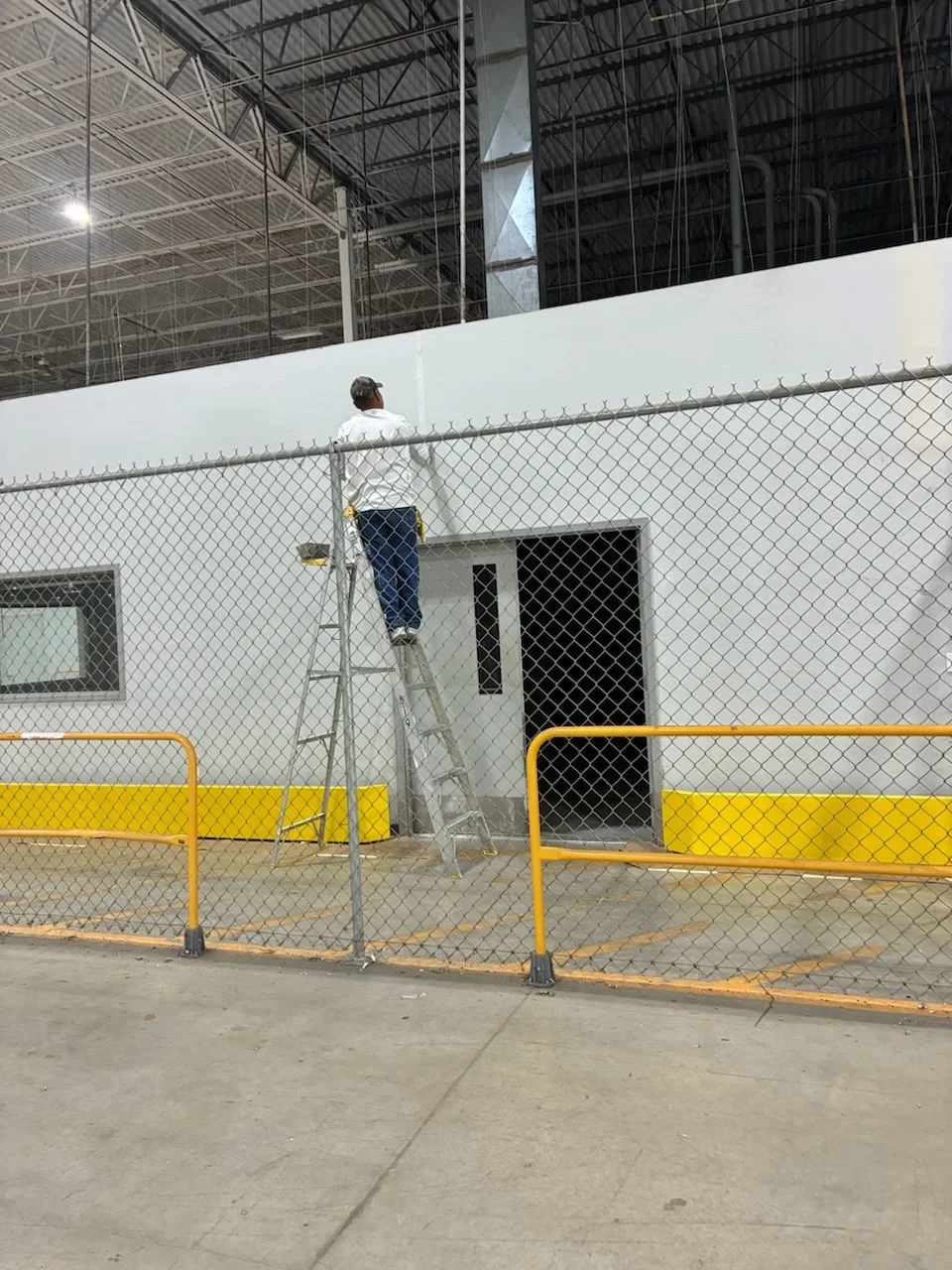 White painted industrial warehouse wall with metal framing, chain-link fence, and yellow safety barriers